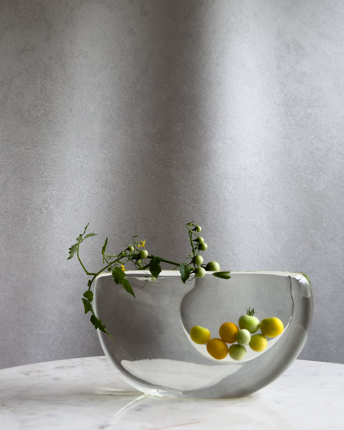 A clear glass bowl-shaped vase with a large cutout, displayed on a white marble surface against a textured gray wall. Inside the vase, yellow and green cherry tomatoes are growing, and a tomato vine with green leaves and small unripe tomatoes is draped over the rim of the vase.