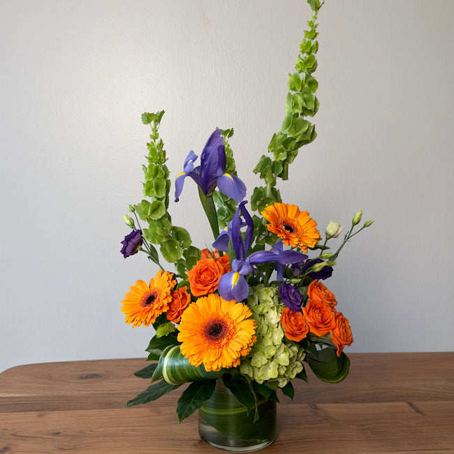 Floral arrangement with orange and purple flowers and green leaves in a clear vase on a wooden surface.