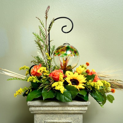 A round glass ornament with a colorful tree design hangs above sunflowers, orange roses, yellow flowers, and greenery.
