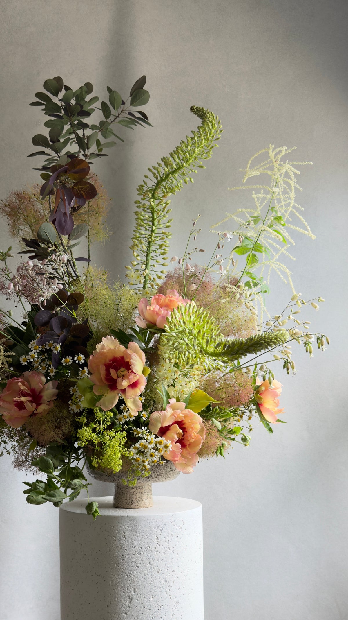 A rustic dried flower arrangement in a textured ceramic pot, displayed on a white cylindrical pedestal against a neutral gray background.
