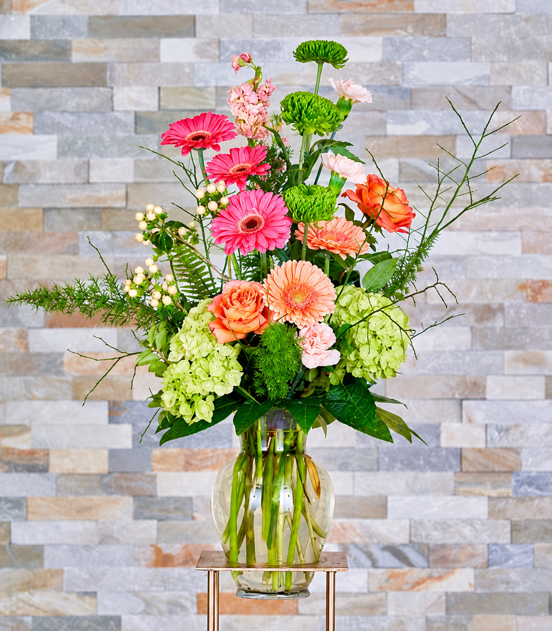 Pink gerberas, peach and orange roses, green hydrangeas and chrysanthemums, and bare branches in a clear glass vase.