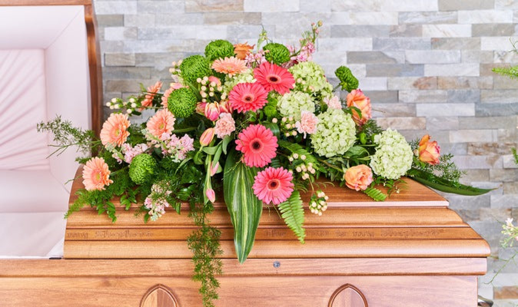 Pink gerberas, peach and orange roses, pale green hydrangeas, green chrysanthemums, and trailing foliage arranged across a wooden casket.