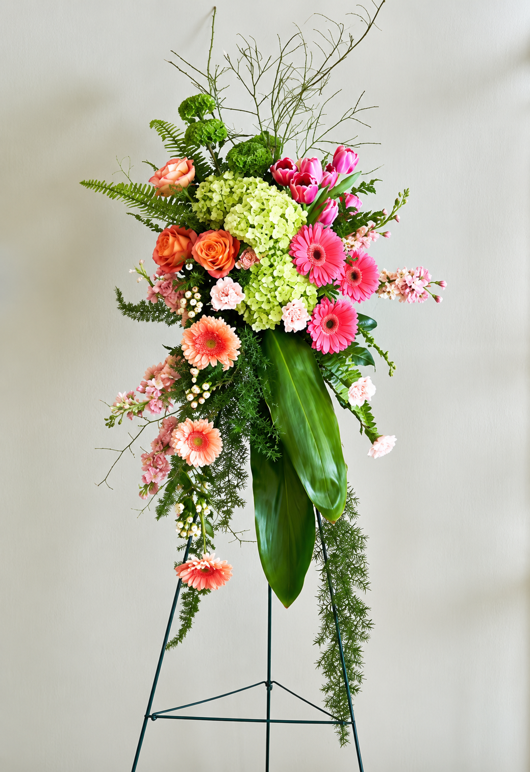Asymmetrical standing spray on an easel with pink gerberas, peach and orange roses, green hydrangeas, bare branches, and long trailing foliage.