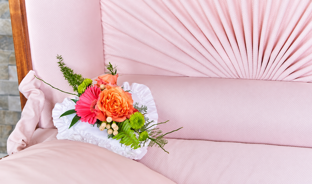 White ruffled casket pillow with a pink gerbera, orange rose, green chrysanthemums, cream berries, and fine greenery against pink casket lining.