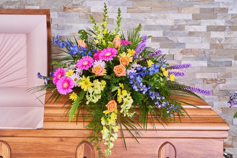 Pink gerberas, peach roses, yellow flowers, blue delphinium, and purple flowering stems with greenery arranged across a wooden casket.