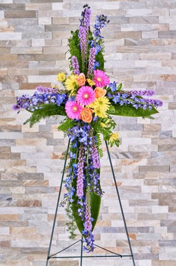 An easel-mounted floral cross with pink gerberas, peach roses, yellow flowers, and long blue and purple flowering stems over greenery.