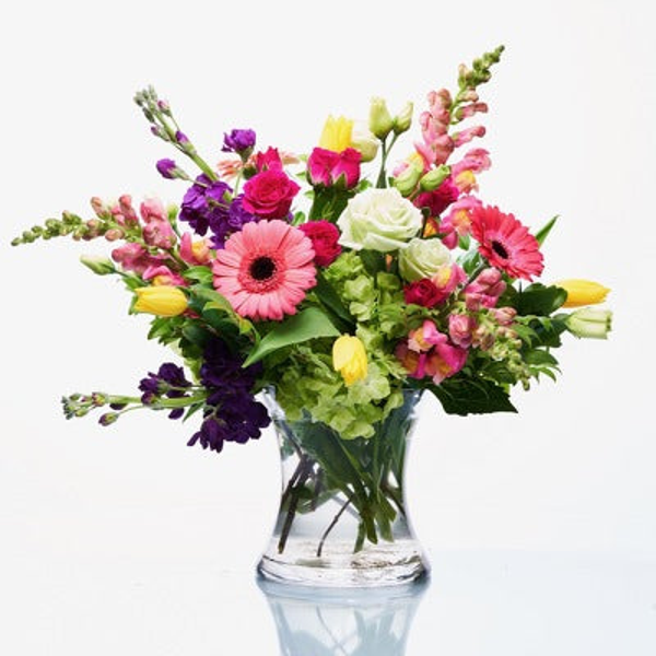 Pink gerberas and roses, white roses, yellow flowers, purple blooms, green hydrangeas, and tall pink flowering stems in a clear glass vase.