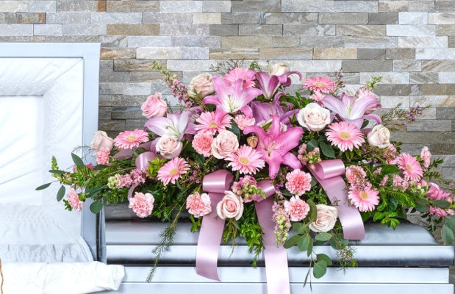 Pink lilies, pale pink roses, pink gerberas and carnations, eucalyptus, greenery, and pink ribbon arranged across a silver casket.