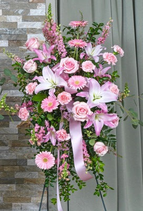Pink lilies, pale pink roses, gerberas, smaller pink flowers, eucalyptus, and long pink ribbon in a standing easel spray.