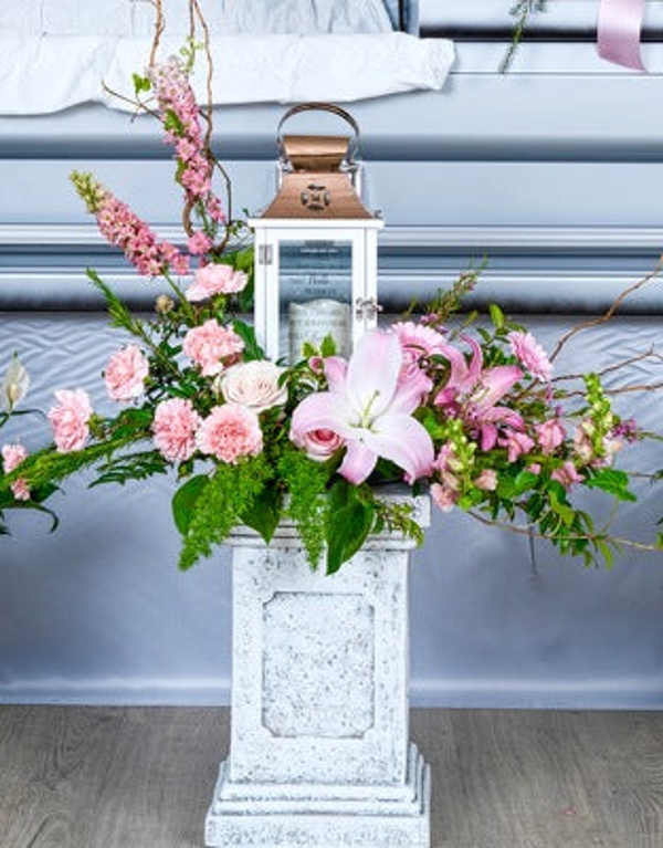 White lantern with a copper-colored top surrounded by pink lilies, pale pink roses, carnations, tall pink flowering stems, bare branches, and ferns.