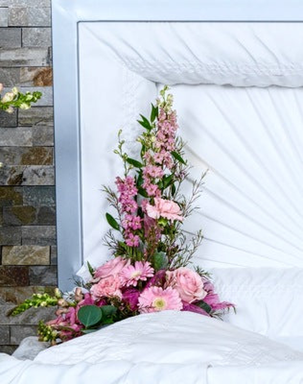 Pale pink roses and gerberas with tall pink flowering stems, eucalyptus, and fine greenery inside a white-lined casket lid.
