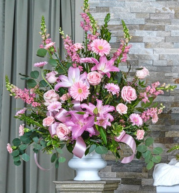 Pink lilies, pale pink roses and gerberas, tall pink flowering stems, eucalyptus, and pink ribbon in a white footed urn-shaped vase.