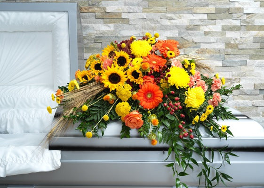 Yellow sunflowers and chrysanthemums, orange gerberas and roses, peach and burgundy flowers, wheat, and trailing greenery across a casket.