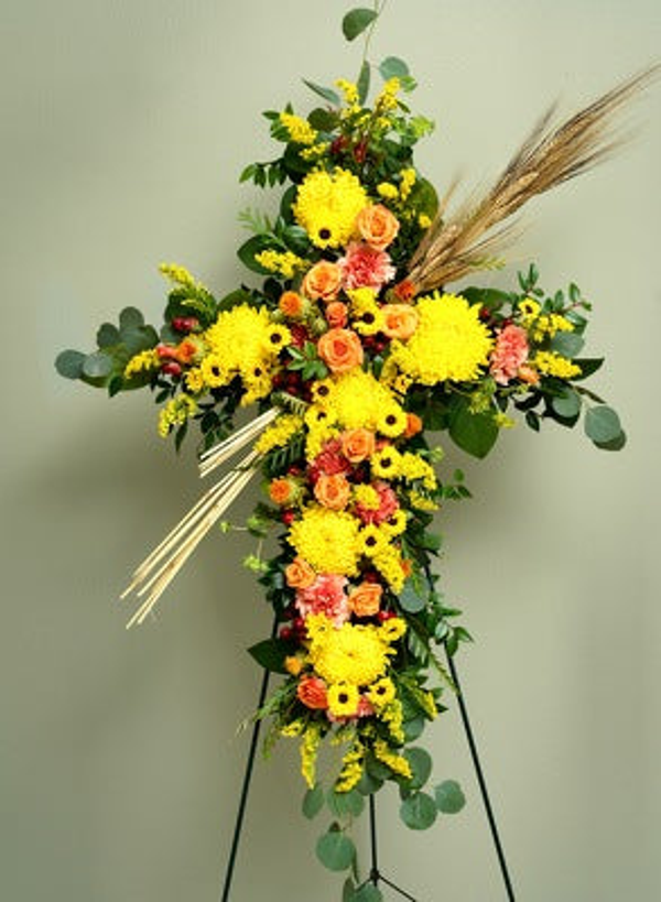 Floral cross on a black easel with yellow chrysanthemums, small sunflowers, orange roses, peach flowers, eucalyptus, and diagonal wheat stems.