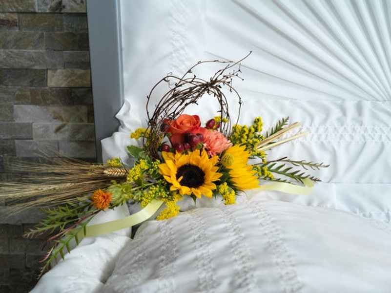 Yellow sunflower, orange and peach roses, burgundy accents, looped branches, wheat, greenery, and pale green ribbon inside a casket lid.