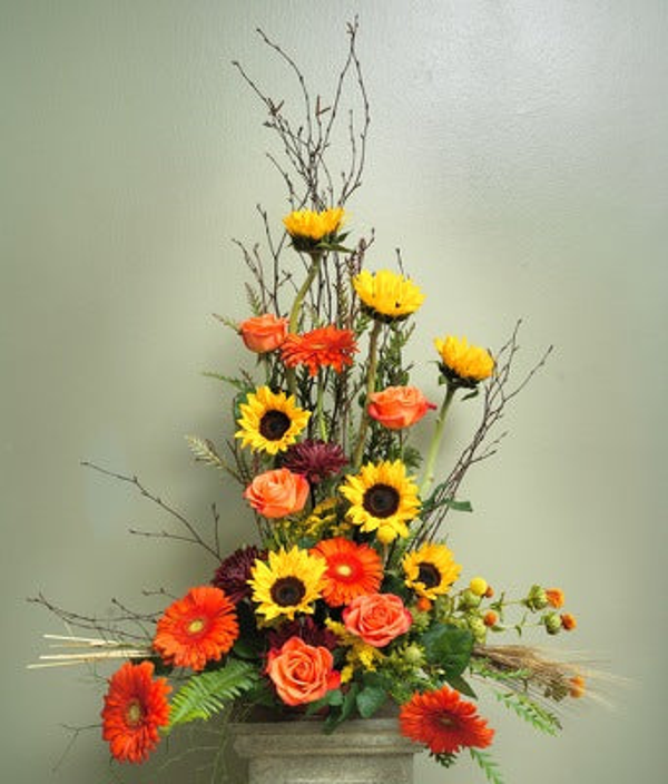 Tall floral tribute with yellow sunflowers and chrysanthemums, orange roses and gerberas, burgundy flowers, bare branches, wheat, and ferns in a low rectangular container.