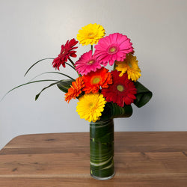 Pink, red, orange, and yellow gerberas at varied heights with curved grasses in a slender cylinder vase lined with green foliage.