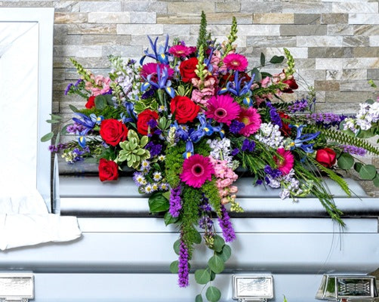 Red roses, deep pink gerberas, blue irises, purple flowering stems, green hydrangeas, and trailing greenery arranged across a casket.
