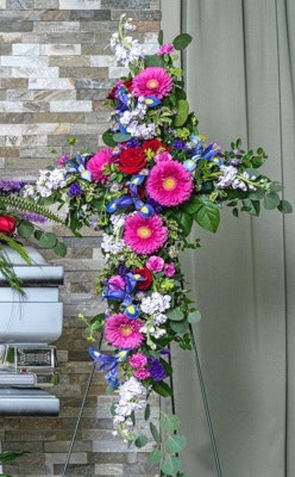 Deep pink gerberas, red roses, blue and purple flowers, small white blooms, and eucalyptus arranged along a cross on an easel.
