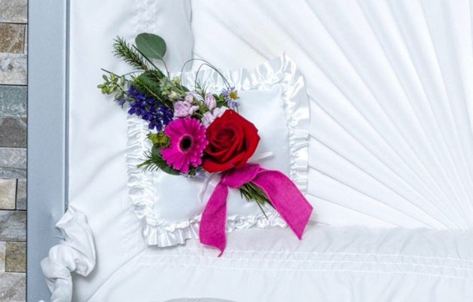 A white ruffled casket pillow with a red rose, deep pink gerbera, blue and purple flowers, greenery, looped grasses, and pink ribbon.