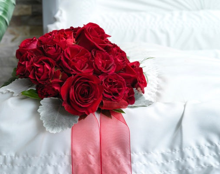 Rounded red rose nosegay edged with silver gray leaves and long pink ribbon inside a white-lined casket.