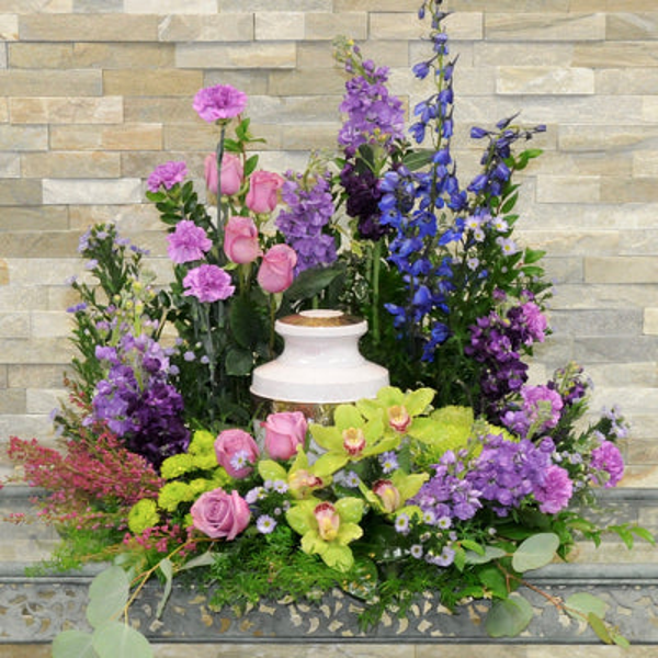 Pink roses, green orchids and chrysanthemums, tall blue and purple flowers, and eucalyptus arranged around a white cremation urn.