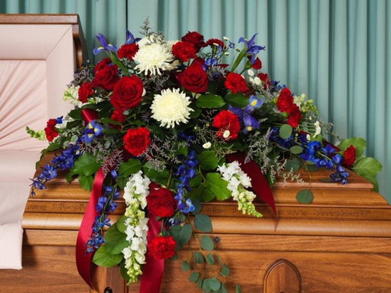Red roses and carnations, white chrysanthemums, blue flowers, trailing greenery, and red ribbon arranged across a wooden casket.
