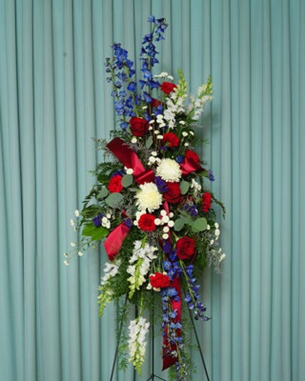 Red roses and carnations, white chrysanthemums, blue delphinium, white trailing flowers, greenery, and red ribbon in a standing easel spray.