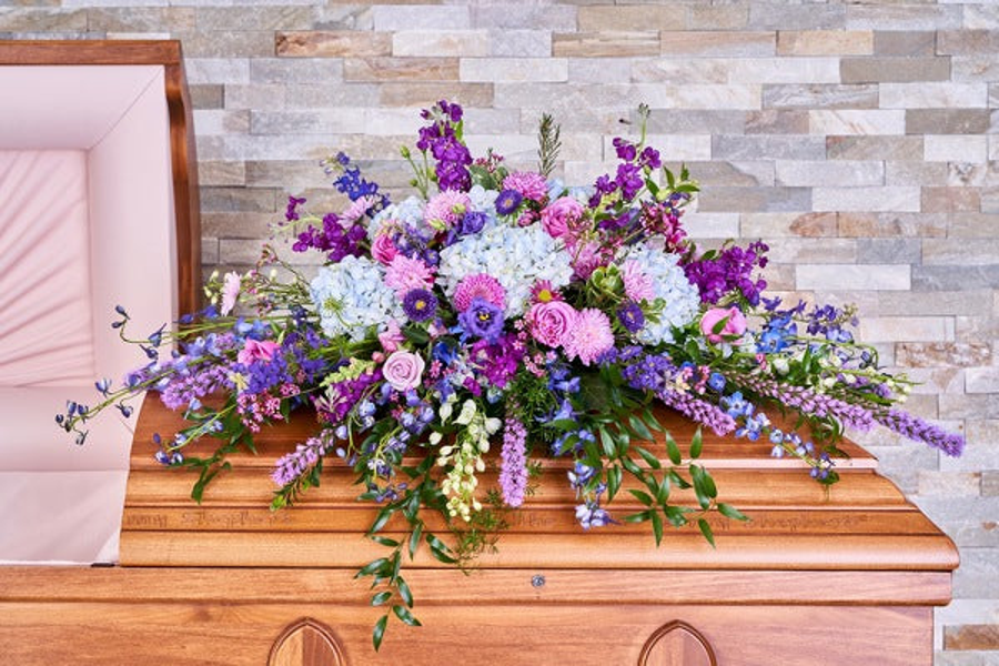 Blue hydrangeas, pink roses, and lavender and purple flowers with tall flowering stems and trailing greenery across a wooden casket.