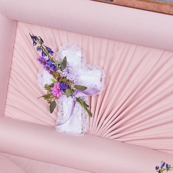White lace-edged cross pillow with blue, pink, and purple flowers, greenery, and lavender ribbon inside a pink-lined casket lid.