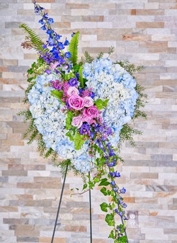 Open heart of pale blue hydrangeas with pink roses, blue and purple flowering stems, ferns, and trailing greenery on an easel.