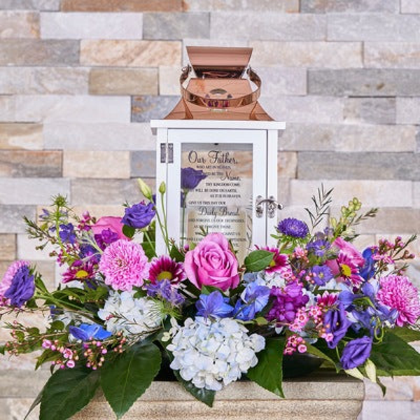 White lantern with a copper-colored top surrounded by pink roses, blue hydrangeas, purple flowers, and spreading greenery.
