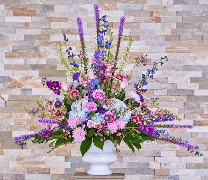 Pink roses, blue hydrangeas, and tall blue and purple flowering stems with ferns and spreading greenery in a white footed urn-shaped vase.