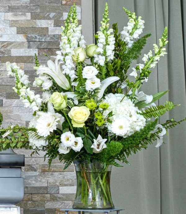 White lilies, pale green roses, white gerberas, and tall white flowering stems with ferns in a clear glass vase.