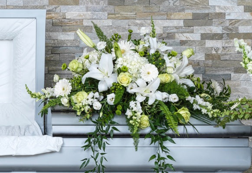 White lilies, hydrangeas, chrysanthemums, and pale green roses with long white flowering stems and trailing greenery across a casket.