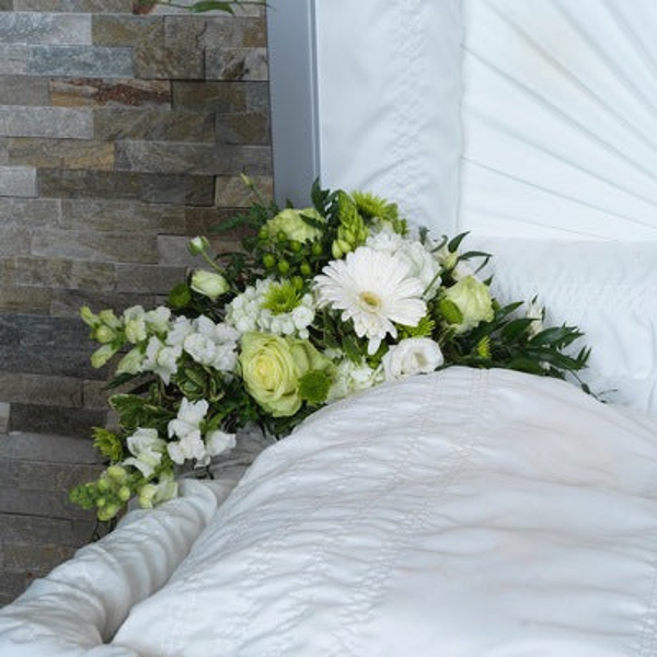 White gerbera, pale green roses, smaller white flowers, and greenery arranged inside an open casket lid.